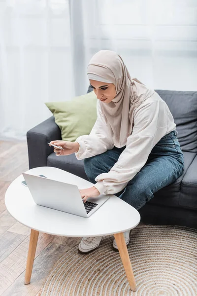 Mujer árabe en hijab usando el ordenador portátil mientras está sentado en el sofá con tarjeta de crédito - foto de stock