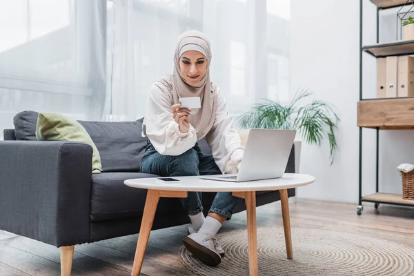 Lächelnde arabische Frau schaut auf Kreditkarte, während sie auf dem Sofa neben dem Laptop sitzt — Stockfoto