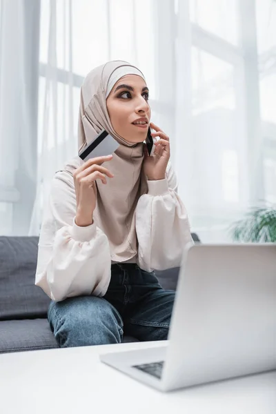 Arabe femme tenant carte de crédit tout en parlant sur téléphone mobile près d'un ordinateur portable — Photo de stock