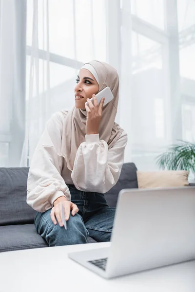 Donna araba sorridente con carta di credito fare ordine su smartphone mentre seduto vicino al computer portatile — Foto stock