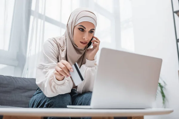 Arabian woman with credit card talking on smartphone near laptop — Stock Photo