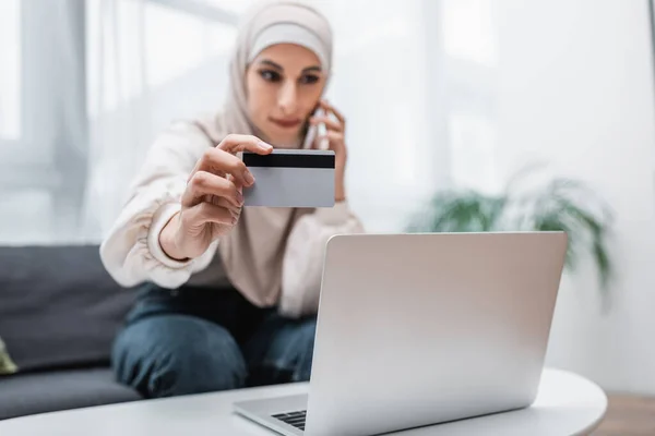 Messa a fuoco selettiva della carta di credito in mano della donna araba offuscata che chiama sullo smartphone vicino al computer portatile — Foto stock