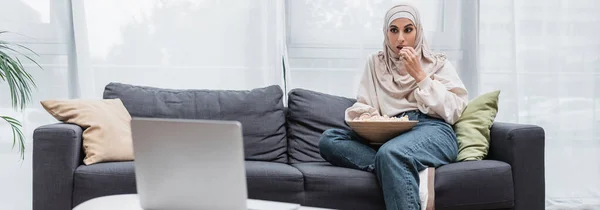 Mujer árabe en hijab comiendo palomitas de maíz y viendo la película en el ordenador portátil en casa, pancarta - foto de stock