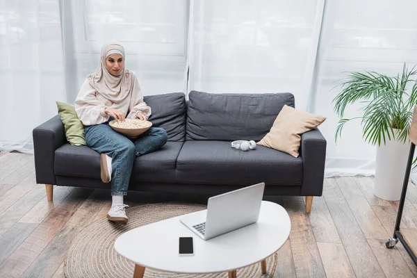 Lächelnde Muslimin schaut Film auf Laptop und isst Popcorn im Wohnzimmer — Stockfoto