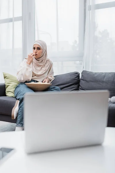 Femme musulmane concentrée manger du pop-corn tout en regardant un film sur ordinateur portable flou — Photo de stock