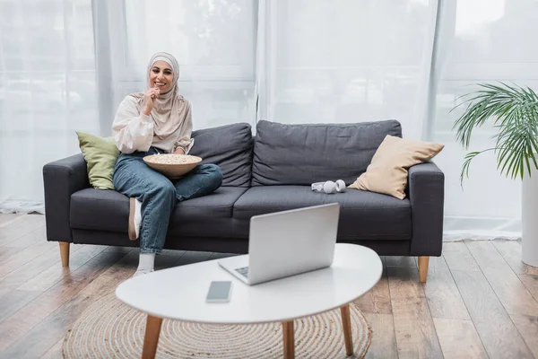 Muslimische Frau mit Schale Popcorn lächelt, während sie im Wohnzimmer einen Film auf dem Laptop anschaut — Stockfoto