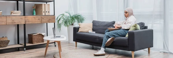 Vue complète de la jeune femme musulmane regardant un film sur ordinateur dans le salon moderne, bannière — Photo de stock