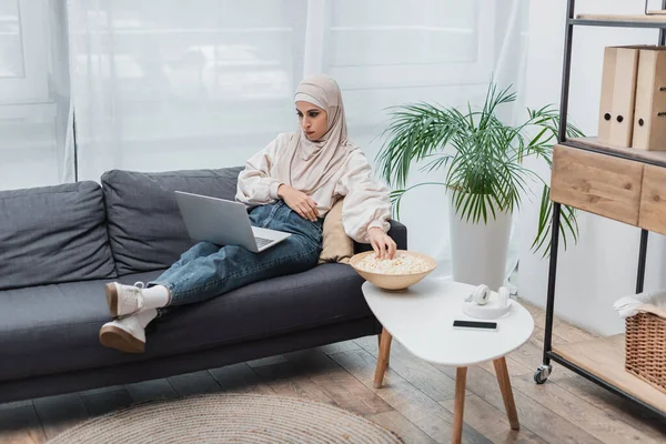 Araba donna guardando film sul computer portatile vicino popcorn e smartphone sul tavolino — Foto stock