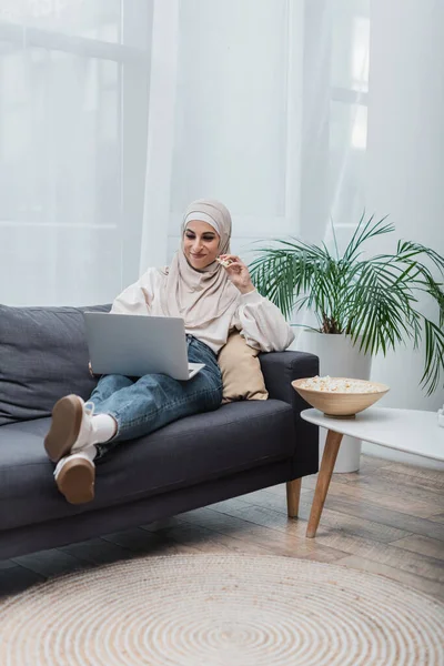 Веселая арабская женщина в хиджабе ест попкорн и смотрит фильм на ноутбуке на диване — стоковое фото