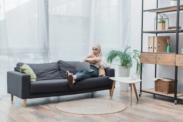 Vue complète de la femme musulmane assise sur le canapé dans le salon moderne et regarder un film sur ordinateur portable — Stock Photo