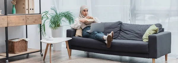 Mujer musulmana sentada con palomitas de maíz cerca de la palma en maceta y viendo la película en el ordenador portátil en la sala de estar, pancarta - foto de stock