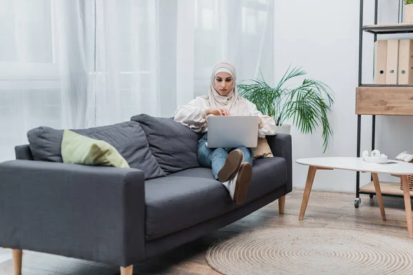 Ganzheitliche Ansicht einer jungen muslimischen Frau, die auf der Couch im Wohnzimmer sitzt und sich den Film auf dem Laptop ansieht — Stockfoto