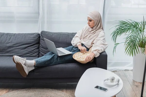 Mulsim mujer en ropa casual y hijab viendo película en el ordenador portátil en la sala de estar - foto de stock