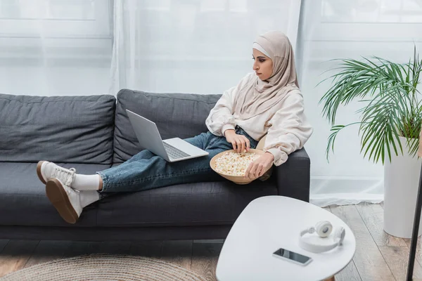 Mulsim woman in casual clothes and hijab watching film on laptop in living room — Stock Photo