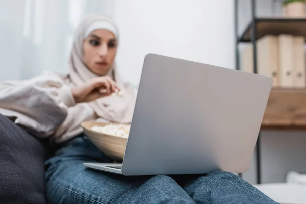 Enfoque selectivo de la computadora portátil cerca de la mujer musulmana borrosa viendo la película en casa - foto de stock