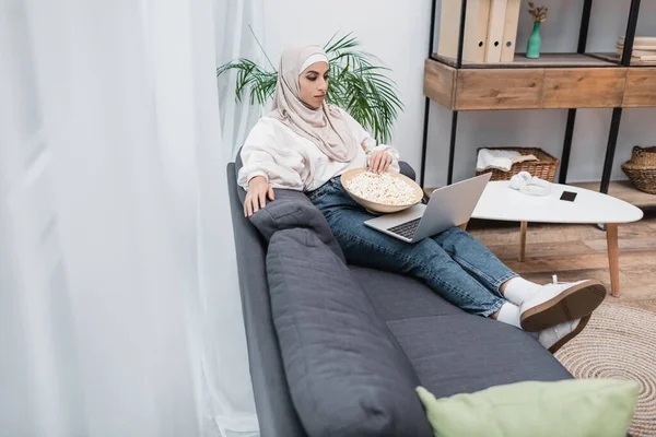 Araberin im Hijab sitzt auf Sofa mit Popcorn und schaut Film am Computer — Stockfoto