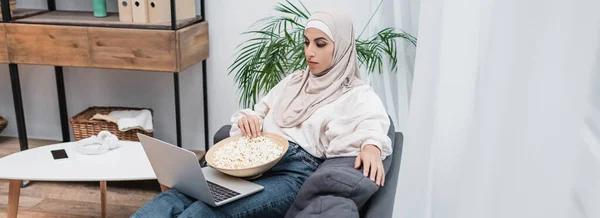 Мусульманська жінка дивиться фільм на ноутбуці біля смартфона з порожнім екраном на кавовому столі, банер — стокове фото