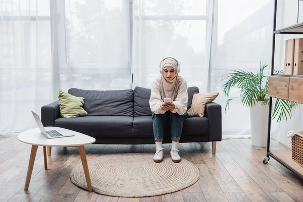 Muslim woman in headphones using smartphone while listening music in living room — Stock Photo