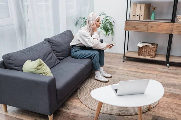 Araberin mit Smartphone hört Musik über Kopfhörer neben Laptop im modernen Wohnzimmer — Stockfoto