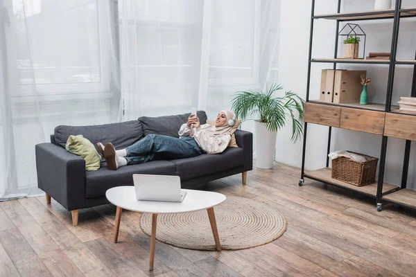 Happy muslim woman listening music in headphones while lying on couch in living room — Stock Photo