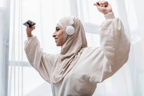 Cheerful muslim woman in headphones holding smartphone while dancing with raised hands — Stock Photo