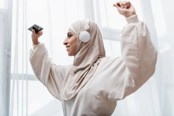 Alegre mujer musulmana en auriculares con teléfono inteligente mientras baila con las manos levantadas - foto de stock
