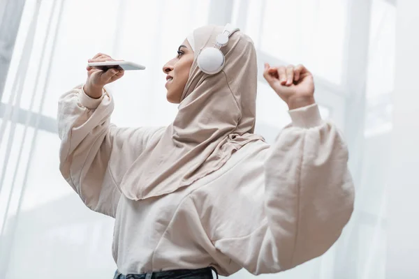 Happy arabian woman in hijab and headphones holding smartphone while dancing at home — Stock Photo