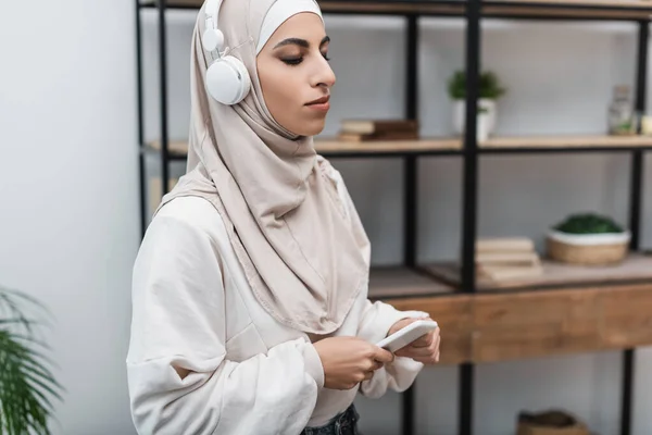 Mujer musulmana escuchando música en auriculares mientras está de pie con el teléfono inteligente en la sala de estar - foto de stock