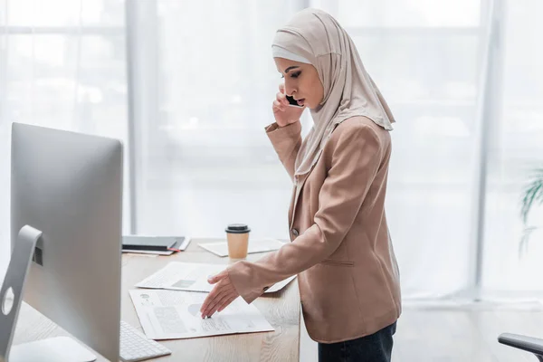 Visão lateral da mulher muçulmana apontando para gráficos enquanto fala no celular perto do monitor do computador — Fotografia de Stock
