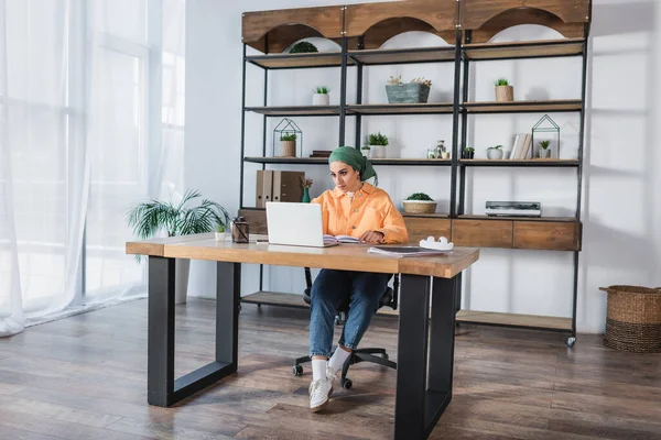 Vista completa de la joven árabe sentada cerca de la computadora portátil mientras estudia en casa - foto de stock