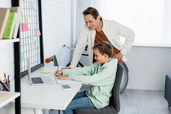 Smiling father standing near son writing on notebook near devices at home — Stock Photo