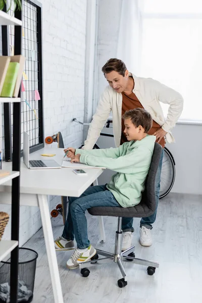 Sonriente niño escribiendo en un cuaderno cerca de dispositivos y papá en casa - foto de stock