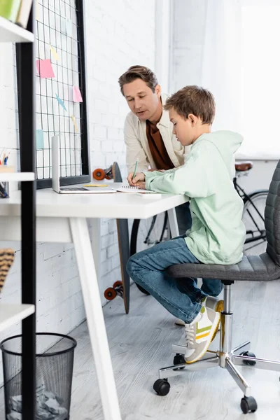 Niño preadolescente escribiendo en un cuaderno cerca de la computadora portátil y papá en casa - foto de stock