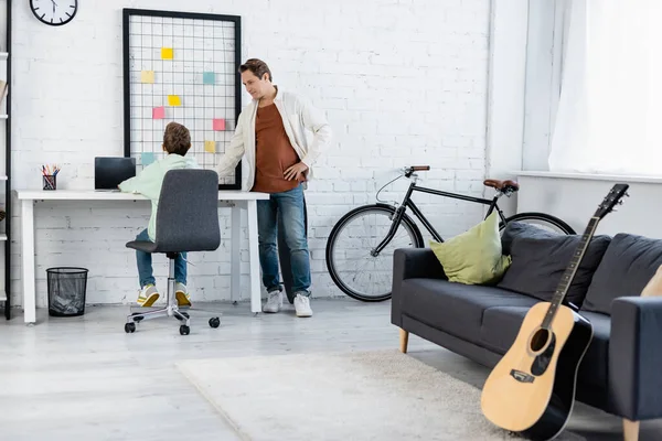 Hombre de pie cerca de hijo usando el ordenador portátil en la sala de estar - foto de stock