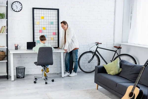 Hombre positivo mirando hijo cerca de la computadora portátil con pantalla en blanco en casa - foto de stock