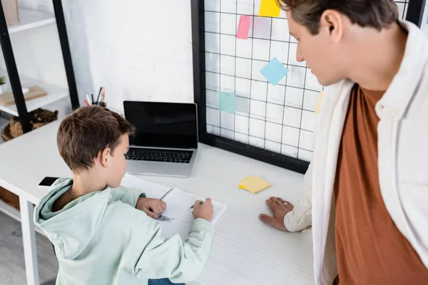 Padre borroso de pie cerca de hijo escribiendo en un cuaderno cerca de dispositivos en casa - foto de stock