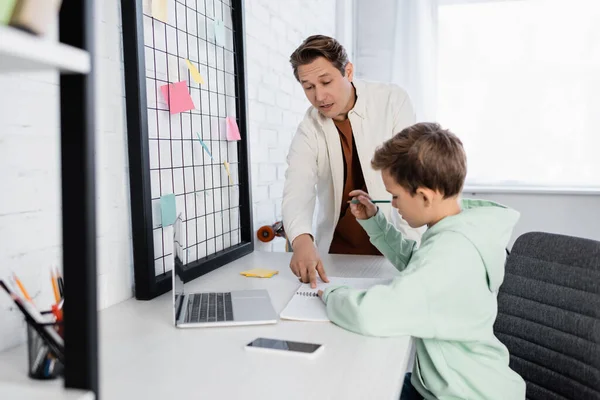 Hombre señalando el cuaderno cerca de hijo y gadgets haciendo la tarea en la sala de estar - foto de stock