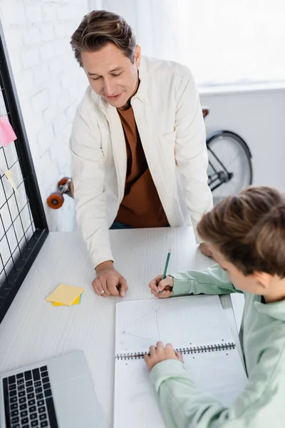 High angle view of smiling man standing near blurred son doing homework in living room — Stock Photo