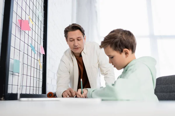 Homme pointant du doigt près de son fils avec crayon et ordinateur portable à la maison — Photo de stock