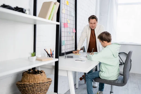 Uomo in piedi vicino a bambino che scrive su notebook vicino a gadget a casa — Foto stock