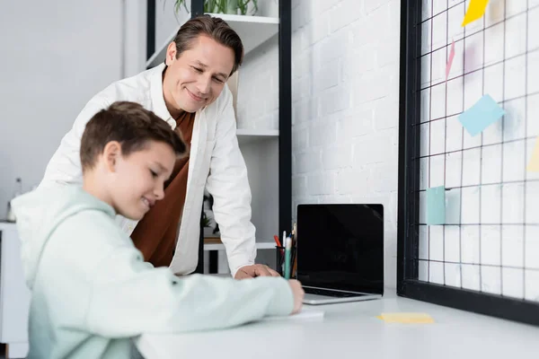 Homem positivo olhando para filho desfocado escrevendo no notebook perto de laptop com tela em branco em casa — Fotografia de Stock