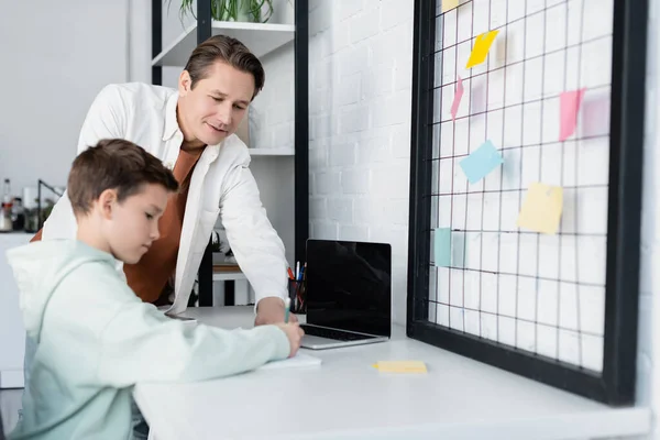 Man standing near son doing homework near laptop and sticky notes at home — Stock Photo