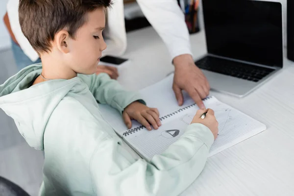 Niño preadolescente escribiendo en el cuaderno cerca de padre y portátil en casa - foto de stock