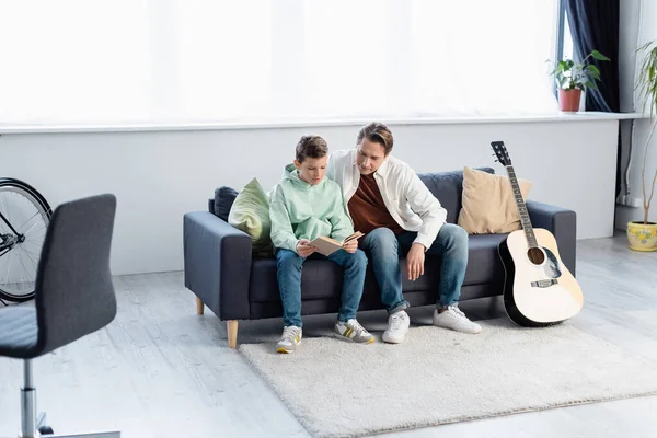 Boy reading book near father on couch in living room — Stock Photo