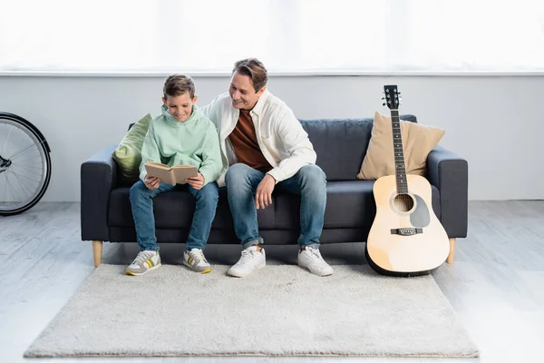 Livre de lecture père et fils près de la guitare acoustique à la maison — Photo de stock