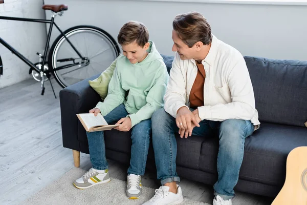 Sonriendo niño preadolescente libro de lectura cerca de papá en el sofá - foto de stock