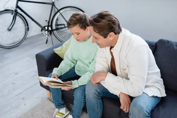 Lächelnder Mann sitzt neben Sohn und liest Buch auf Sofa im Wohnzimmer — Stockfoto