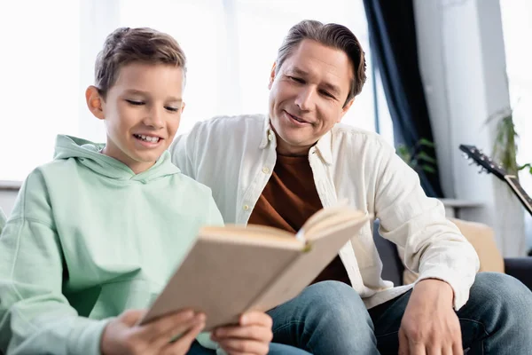 Sonriente chico leyendo libro borroso cerca de papá en casa - foto de stock