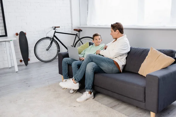 Niño positivo usando el ordenador portátil cerca del padre en el sofá - foto de stock