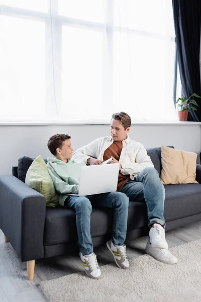 Smiling child using laptop near dad with cellphone on couch — Stock Photo